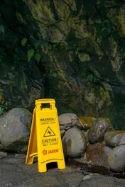 A bright yellow caution sign stands on a wet, rocky path against a mossy stone wall. The sign warns of a slippery floor in both English and Indonesian. Ferns and other greenery grow from the wall, and the stones are dark and wet from moisture.