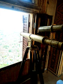 A vintage telescope on a tripod stands near a window in a richly decorated room, providing a view of a cityscape outside. The walls and ceiling have intricate patterns and designs, contributing to an antique and historical atmosphere.