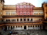 An intricate historical building with a pink and sandstone facade, featuring numerous small windows with latticed designs. The architecture is ornate with traditional Indian motifs, including arches and decorative elements. The structure has multiple levels with repetitive patterns and a prominent central portion.