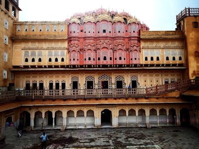 An intricate historical building with a pink and sandstone facade, featuring numerous small windows with latticed designs. The architecture is ornate with traditional Indian motifs, including arches and decorative elements. The structure has multiple levels with repetitive patterns and a prominent central portion.