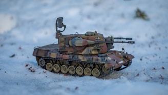 A model of a military tank is positioned on a snow-covered ground. The tank is detailed with a camouflage pattern and features a mounted turret and multiple wheels. Surrounding the tank are small twigs and bits of foliage partially embedded in the snow.
