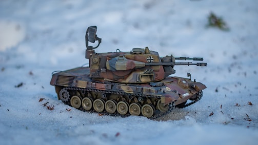 A model of a military tank is positioned on a snow-covered ground. The tank is detailed with a camouflage pattern and features a mounted turret and multiple wheels. Surrounding the tank are small twigs and bits of foliage partially embedded in the snow.