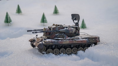 A model tank is positioned on a snowy landscape with miniature evergreen trees in the background. The tank features intricate details, including weaponry and camouflage paint.