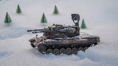 A model tank is positioned on a snowy landscape with miniature evergreen trees in the background. The tank features intricate details, including weaponry and camouflage paint.