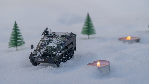 A miniature military vehicle is set on a snowy landscape, surrounded by small lit tealight candles and tiny model pine trees. The scene creates a winter diorama effect.