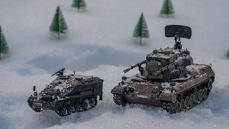 Two miniature military vehicles are placed on a snowy surface, surrounded by small, conical green trees. The scene resembles a winter landscape diorama. The tanks are detailed, with camouflage patterns and equipment mounted on them.