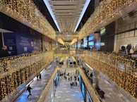 A multi-level shopping mall decorated with strings of yellow lights, viewed from an elevated perspective. Shoppers are walking on the lower level, while upper levels feature storefronts and railings adorned with lights. The marble floor adds a touch of elegance, contributing to a festive atmosphere.