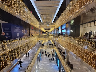 A multi-level shopping mall decorated with strings of yellow lights, viewed from an elevated perspective. Shoppers are walking on the lower level, while upper levels feature storefronts and railings adorned with lights. The marble floor adds a touch of elegance, contributing to a festive atmosphere.