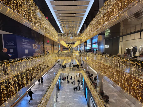 A multi-level shopping mall decorated with strings of yellow lights, viewed from an elevated perspective. Shoppers are walking on the lower level, while upper levels feature storefronts and railings adorned with lights. The marble floor adds a touch of elegance, contributing to a festive atmosphere.