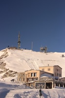 A snowy mountain landscape with several buildings near the base, equipped with antenna towers for communication. The sky above is clear and bright blue. Tracks in the snow suggest recent skiing or snowboarding activity.