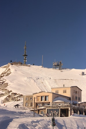 A snowy mountain landscape with several buildings near the base, equipped with antenna towers for communication. The sky above is clear and bright blue. Tracks in the snow suggest recent skiing or snowboarding activity.