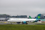 An Aer Lingus airplane is taxiing on the runway at an airport. The background shows terminal buildings with other aircraft parked nearby. The sky is overcast, creating a cloudy atmosphere.