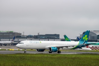 An Aer Lingus airplane is taxiing on the runway at an airport. The background shows terminal buildings with other aircraft parked nearby. The sky is overcast, creating a cloudy atmosphere.