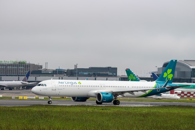 An Aer Lingus airplane is taxiing on the runway at an airport. The background shows terminal buildings with other aircraft parked nearby. The sky is overcast, creating a cloudy atmosphere.