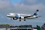 An airplane with white and blue colors is captured mid-air with its landing gear extended, flying against a cloudy sky. The aircraft has 'EGYPTAIR' written prominently on its fuselage, and it features a logo on the tail. In the background, an airport with various terminal buildings and other parked airplanes can be seen.