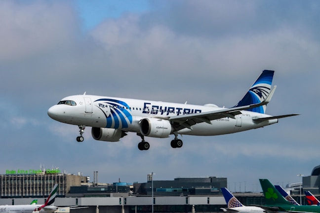 An airplane with white and blue colors is captured mid-air with its landing gear extended, flying against a cloudy sky. The aircraft has 'EGYPTAIR' written prominently on its fuselage, and it features a logo on the tail. In the background, an airport with various terminal buildings and other parked airplanes can be seen.