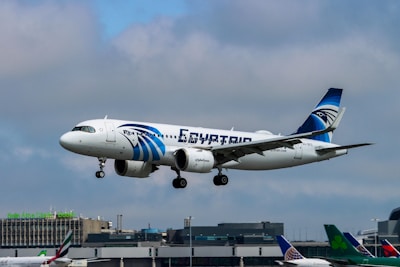An airplane with white and blue colors is captured mid-air with its landing gear extended, flying against a cloudy sky. The aircraft has 'EGYPTAIR' written prominently on its fuselage, and it features a logo on the tail. In the background, an airport with various terminal buildings and other parked airplanes can be seen.