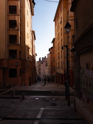 A narrow street between tall, warm-toned buildings under a clear blue sky. The architecture is historic, with balconies and street lamps. Cobblestones cover the street, leading to smaller buildings in the distance.