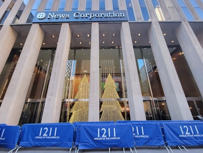 Large glass facade of a building with the 'News Corporation' sign above. Inside, two illuminated Christmas trees are visible. Blue barriers with '1211 Avenue of the Americas' in white text line the front.