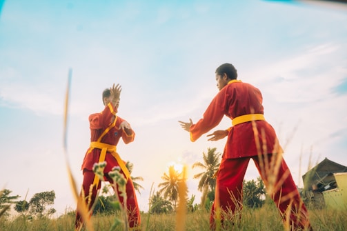 Two individuals dressed in matching red martial arts uniforms with yellow belts are practicing or sparring in a grassy field. The sun is setting or rising in the background, casting a warm glow over the scene. Palm trees are visible, adding an element of tropical scenery.