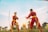 Two individuals dressed in matching red martial arts uniforms with yellow belts are practicing or sparring in a grassy field. The sun is setting or rising in the background, casting a warm glow over the scene. Palm trees are visible, adding an element of tropical scenery.