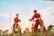 Two individuals dressed in matching red martial arts uniforms with yellow belts are practicing or sparring in a grassy field. The sun is setting or rising in the background, casting a warm glow over the scene. Palm trees are visible, adding an element of tropical scenery.
