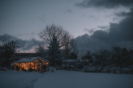 A snow-covered garden is illuminated by a warmly lit greenhouse. The sky is filled with dark clouds and the setting appears serene and wintery with trees and shrubs blanketed in snow.