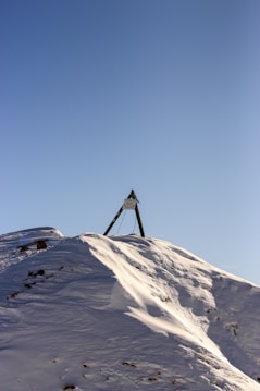 A snow-covered mountain peak with a metal tripod structure at the summit, set against a clear blue sky.