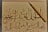 An intricate calligraphy piece featuring Arabic script is displayed on off-white paper. A wooden calligraphy pen is positioned diagonally on the right side of the paper. The calligraphy includes elegant, flowing lines and various diacritical marks.