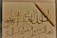 An intricate calligraphy piece featuring Arabic script is displayed on off-white paper. A wooden calligraphy pen is positioned diagonally on the right side of the paper. The calligraphy includes elegant, flowing lines and various diacritical marks.