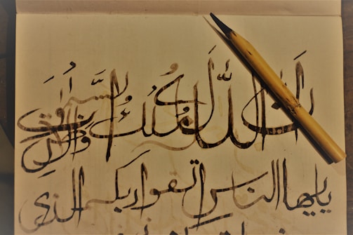 An intricate calligraphy piece featuring Arabic script is displayed on off-white paper. A wooden calligraphy pen is positioned diagonally on the right side of the paper. The calligraphy includes elegant, flowing lines and various diacritical marks.