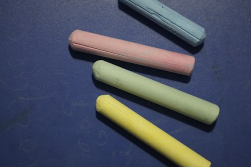 Four pieces of colored chalk are arranged on a dark blue surface. The chalks are in pastel shades of blue, pink, green, and yellow, creating a soft and playful contrast against the background.