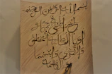 A close-up of a hand writing Urdu poetry on textured paper under soft, moody lighting.