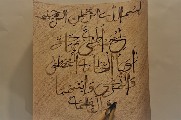 A piece of paper with Arabic calligraphy written in black ink is placed on a textured, light brown background. A wooden pen is lying at the bottom right corner of the paper, partially visible.