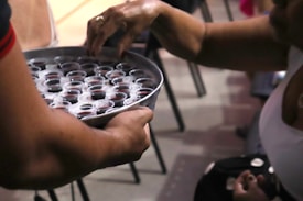 A person is holding a tray filled with small cups containing a dark liquid. Another hand reaches to pick up one of the cups. The scene suggests a communal setting, possibly involving a shared activity or ritual.