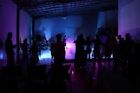 Silhouettes of people are gathered in a dimly lit room, with colorful lighting in shades of purple, pink, and blue creating an ambient atmosphere. Some appear to be dancing or engaged in conversation near a music setup, possibly involving a live performance with instruments.