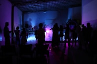 Silhouettes of people are gathered in a dimly lit room, with colorful lighting in shades of purple, pink, and blue creating an ambient atmosphere. Some appear to be dancing or engaged in conversation near a music setup, possibly involving a live performance with instruments.