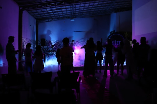 Silhouettes of people are gathered in a dimly lit room, with colorful lighting in shades of purple, pink, and blue creating an ambient atmosphere. Some appear to be dancing or engaged in conversation near a music setup, possibly involving a live performance with instruments.