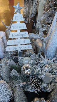 A festive holiday display featuring a white sign shaped like a Christmas tree with silver glittery text, surrounded by silver reindeer, snow-covered pine branches, and various winter-themed ornaments. The scene includes silver and white decorations such as a star on top of the sign and glittering cones.