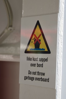 A warning sign affixed to a white wall, featuring a yellow triangle with a black border. Inside the triangle, there is an image of a trash can with garbage and a red cross over it, symbolizing prohibition. Below the triangle, text in both Norwegian and English reads 'Do not throw garbage overboard.'