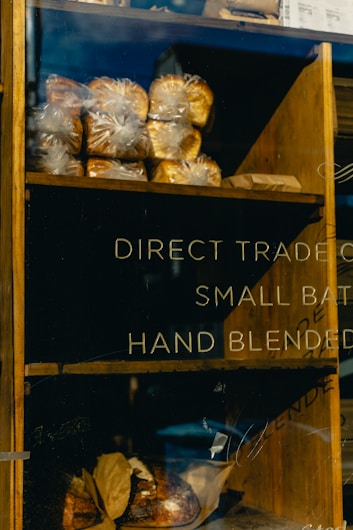 A wooden shelf inside a bakery displays several loaves of bread wrapped in plastic. The shelves are partially filled, with some loaves in the background and others on a lower shelf. Text on the glass window reads 'DIRECT TRADE,' 'SMALL BATCH,' and 'HAND BLENDED,' indicating the bakery's emphasis on artisanal products.