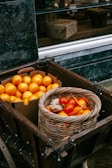 A wooden cart holds a box filled with bright oranges and a wicker basket containing red tomatoes, ginger, and yellow peppers. The scene is set against a dark wall background with a reflective window showing part of a shop interior with a wrapped gift and wicker items.