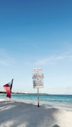 A sandy beach with a signpost featuring several signs pointing towards a restaurant named Aranza. The signs also mention 'ceviches', 'cocteles', and 'pescados', indicating food options. A flag with red, white, and blue colors is planted in the sand nearby. The ocean and clear blue sky create a serene background.