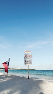 A sandy beach with a signpost featuring several signs pointing towards a restaurant named Aranza. The signs also mention 'ceviches', 'cocteles', and 'pescados', indicating food options. A flag with red, white, and blue colors is planted in the sand nearby. The ocean and clear blue sky create a serene background.