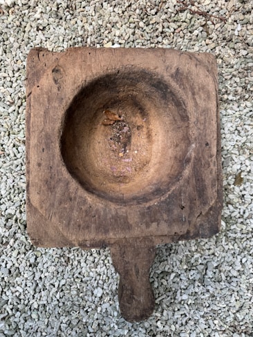 A rustic, weathered wooden bowl with a handle is set on a background of light-colored pebbles. The bowl has a round shallow indentation and appears quite old, with visible cracks and a rough texture. Inside the bowl, there are small remnants of leaves and dirt.