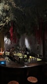 A dimly lit bar with a rustic atmosphere. Overhead, green foliage and hanging red ribbons create a dramatic ceiling display. The bartender is preparing drinks behind a counter filled with bottles and utensils. Warm lighting enhances the cozy ambiance, while patrons sit at the counter.