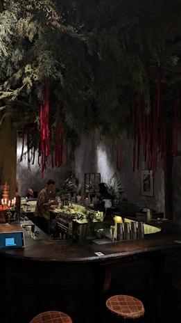 A dimly lit bar with a rustic atmosphere. Overhead, green foliage and hanging red ribbons create a dramatic ceiling display. The bartender is preparing drinks behind a counter filled with bottles and utensils. Warm lighting enhances the cozy ambiance, while patrons sit at the counter.