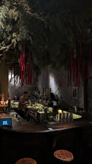 A dimly lit bar with a rustic atmosphere. Overhead, green foliage and hanging red ribbons create a dramatic ceiling display. The bartender is preparing drinks behind a counter filled with bottles and utensils. Warm lighting enhances the cozy ambiance, while patrons sit at the counter.