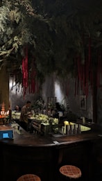 A dimly lit bar with a rustic atmosphere. Overhead, green foliage and hanging red ribbons create a dramatic ceiling display. The bartender is preparing drinks behind a counter filled with bottles and utensils. Warm lighting enhances the cozy ambiance, while patrons sit at the counter.