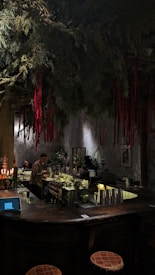 A dimly lit bar with a rustic atmosphere. Overhead, green foliage and hanging red ribbons create a dramatic ceiling display. The bartender is preparing drinks behind a counter filled with bottles and utensils. Warm lighting enhances the cozy ambiance, while patrons sit at the counter.